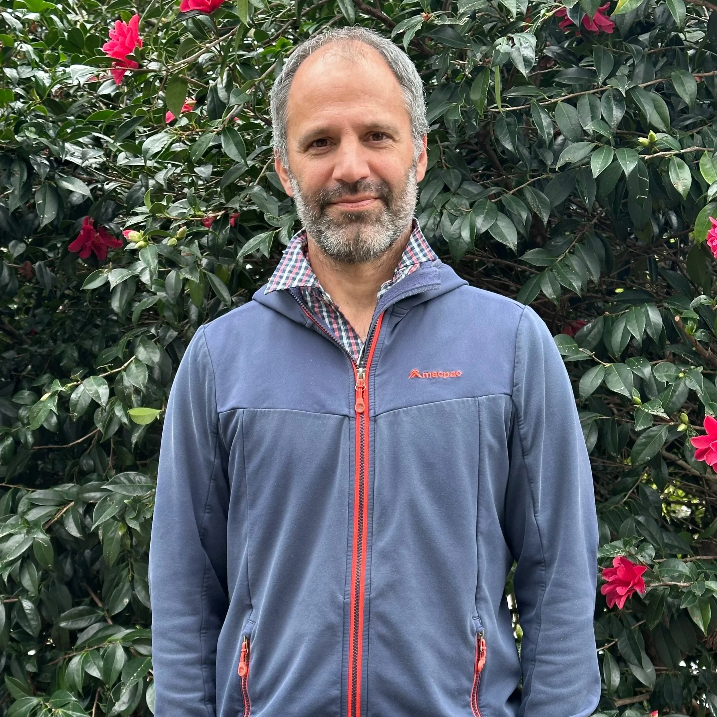 Aaron Agnew smiles at the camera. He is wearing a bluse hoodie with a red zip up the middle.
