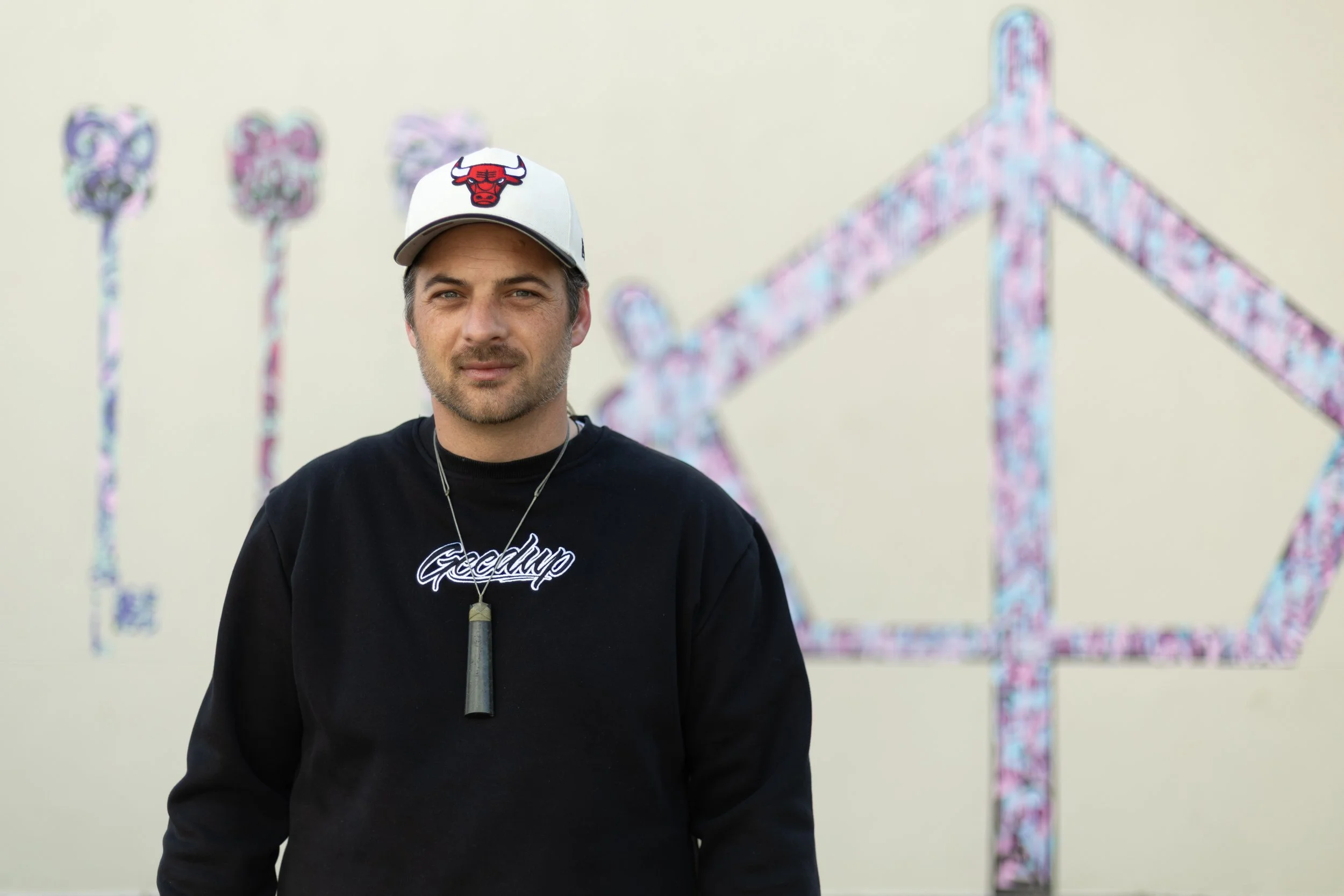 A pic of Logan looking straight down the camera lens. He wears a black sweatshirt with the words Goodup, a chicago bulls cap and a pounamu toki.