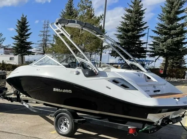 White and black motorboat on trailer in parking lot with trees and blue sky in background.