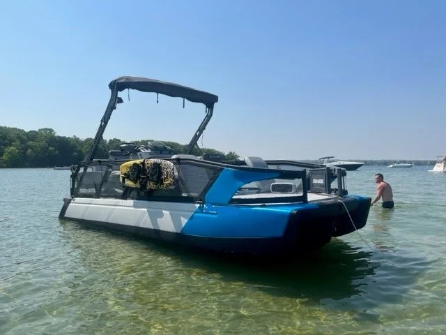 Blue and black pontoon boat anchored in shallow water with a man swimming nearby, trees and other boats in the background.