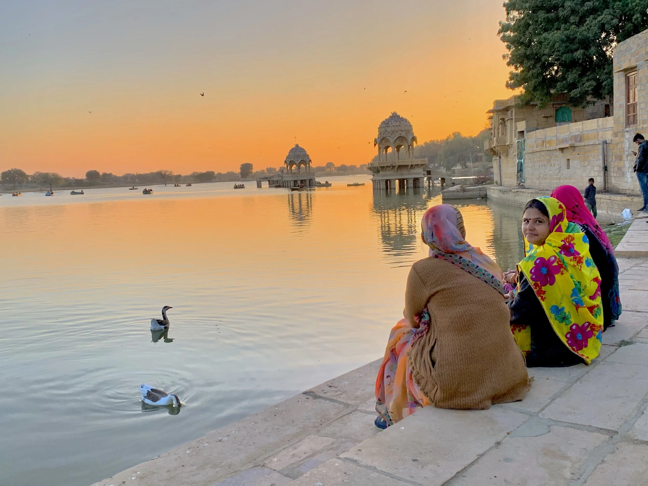 Women and children sitting by a river at sunset, with boats and historic architecture, including pavilions and a stone building, reflected in the water.