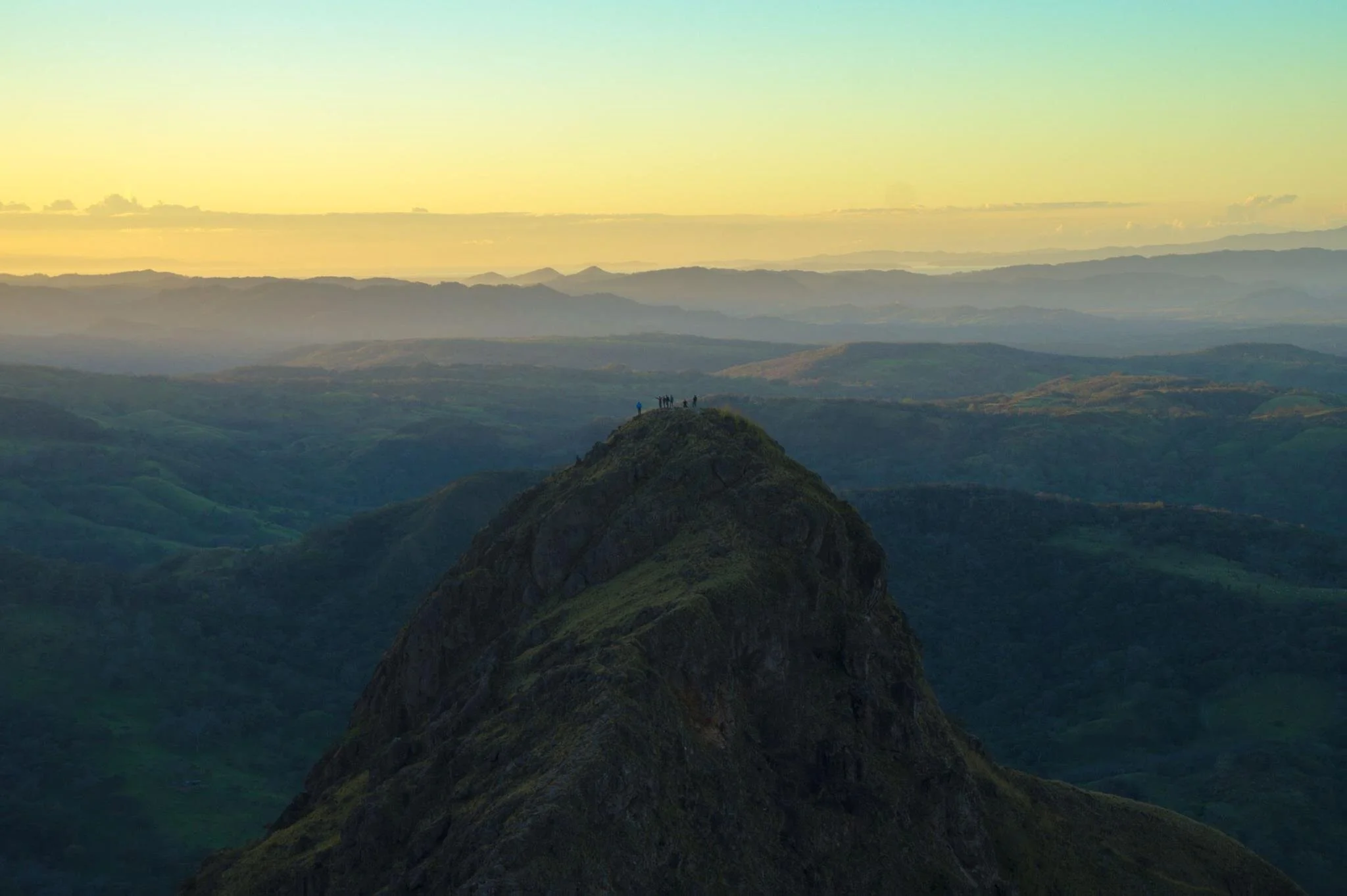 A small group of people standing on a mountain peak during sunrise or sunset, overlooking a series of rolling hills and mountain ranges in the distance.