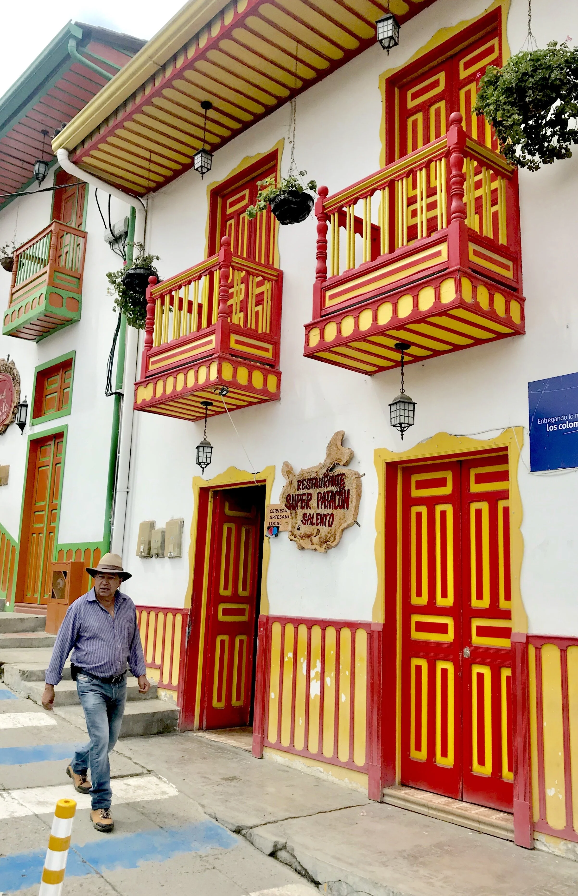 A colorful building with red and yellow accents, featuring balconies and decorative doors. A man in a hat and striped shirt walks on the sidewalk in front of the building.