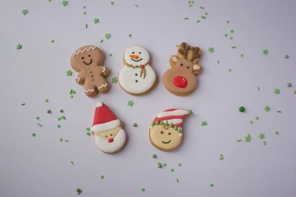 Five decorated holiday cookies on a white surface, including a gingerbread man, snowman, reindeer, Santa Claus, and an elf, with green sprinkles scattered around.