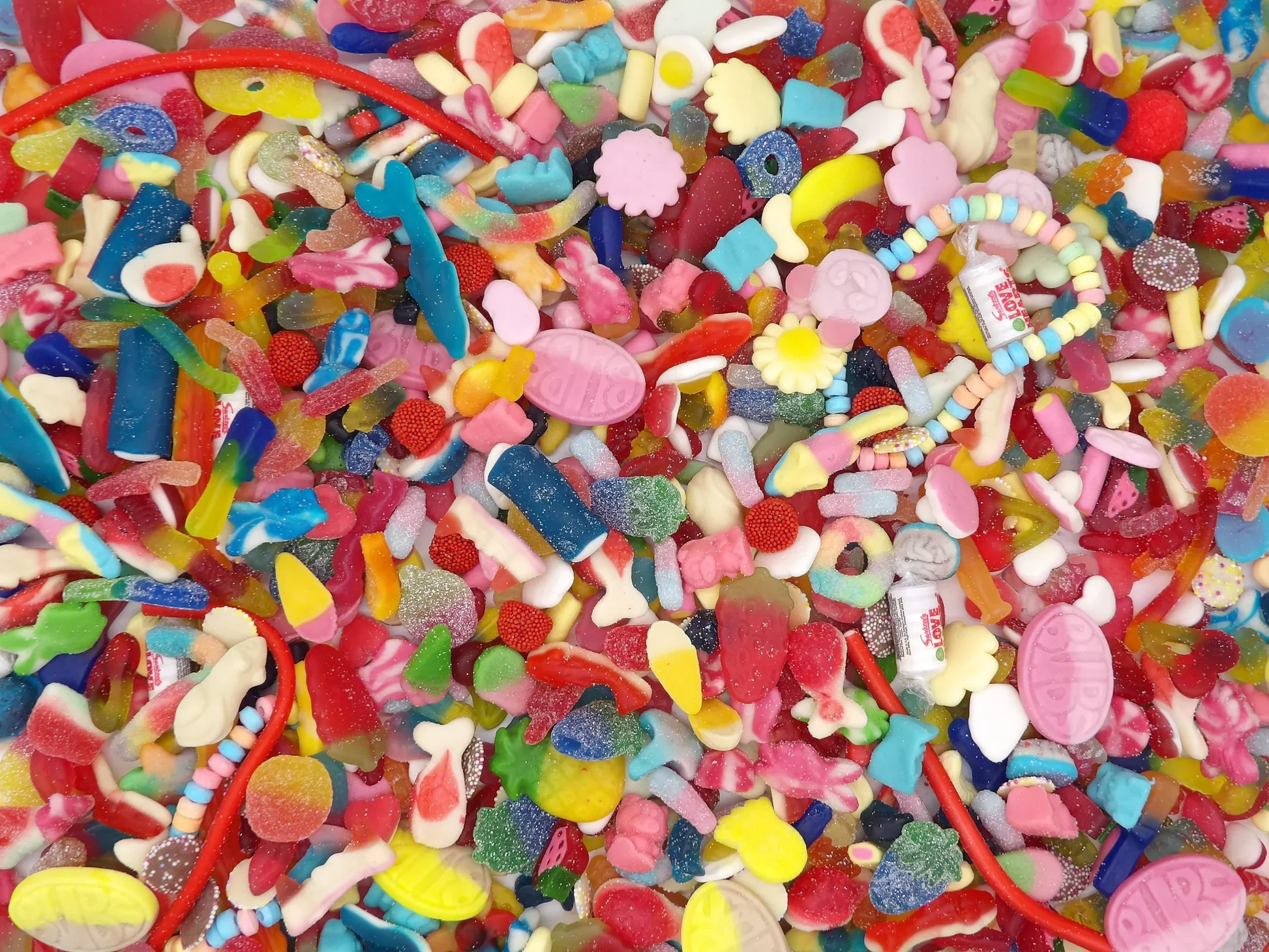 Colorful assortment of various gummy candies, jelly beans, gummy worms, and small candy shapes in bright colors like red, pink, yellow, green, and blue.