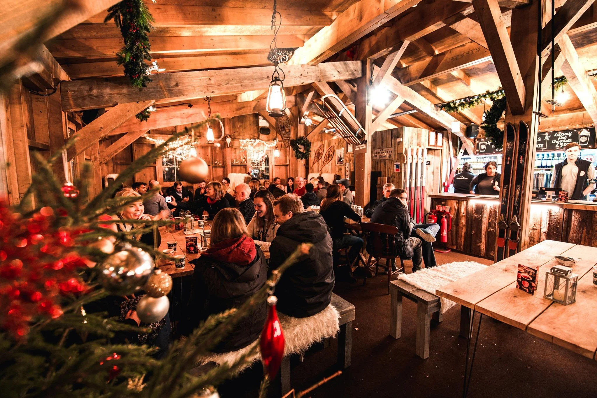 A cozy, festive indoor restaurant decorated for Christmas with a Christmas tree in the foreground, walls and ceiling made of wood, warm lighting, and holiday ornaments. Many people are sitting at tables, enjoying their meals, and socializing.