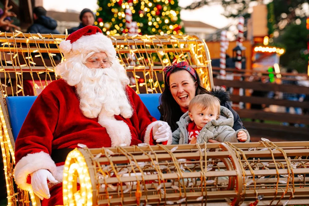 Santa Claus, a woman, and a child enjoying a ride on a festive Christmas float decorated with lights, with a Christmas tree and holiday decorations in the background.