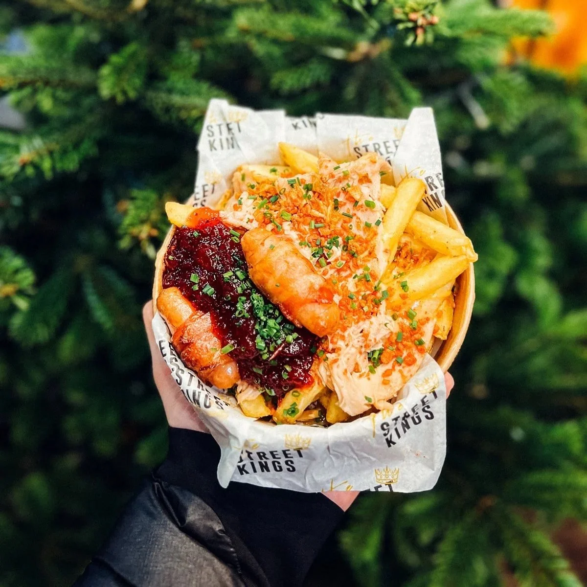 A hand holding a container of loaded fries with chicken, topped with sauce, herbs, and cheese, in front of a Christmas tree.