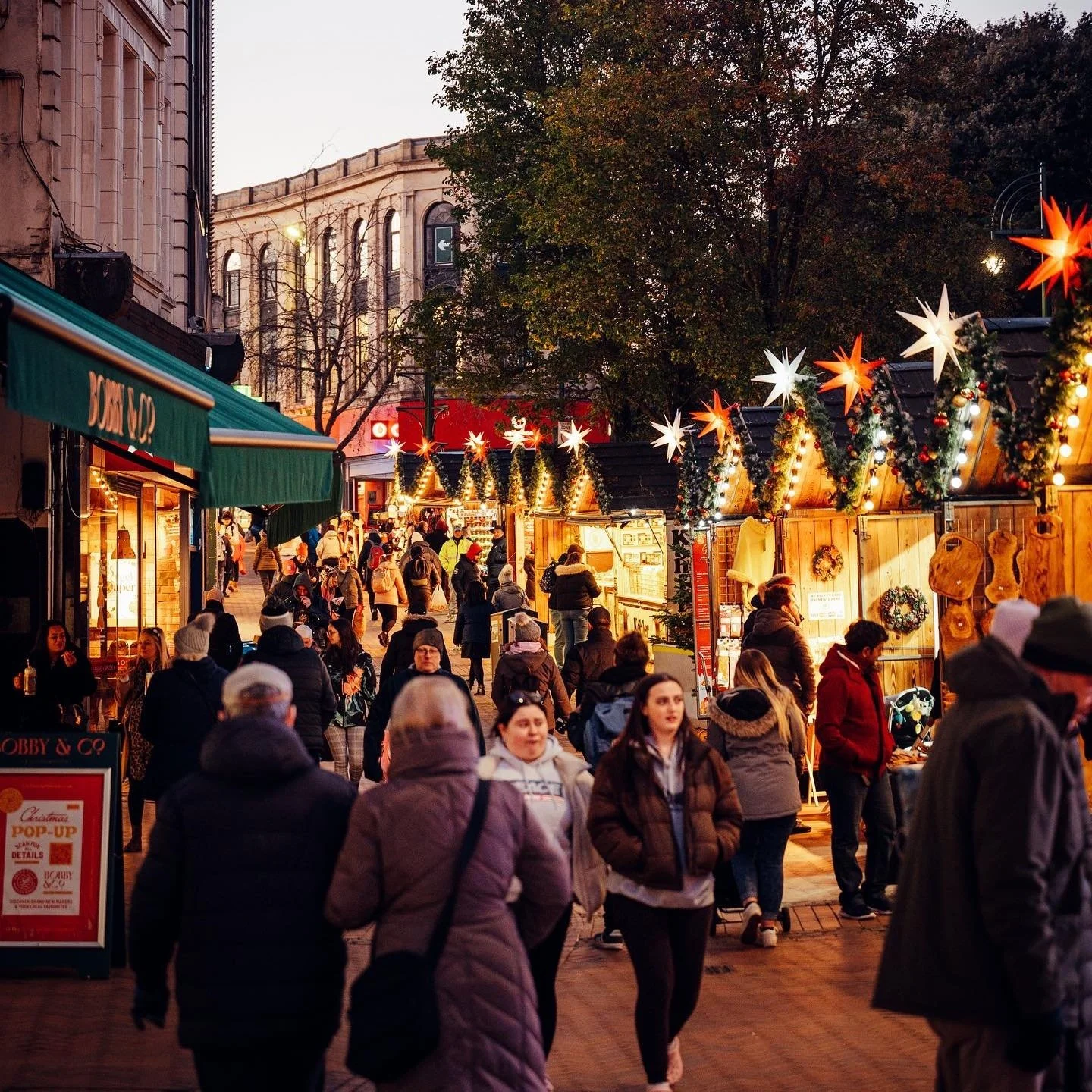 A busy outdoor Christmas market in the evening with wooden stalls decorated with string lights, Christmas wreaths, and star-shaped ornaments. People are walking, shopping, and socializing on the street lined with historic buildings, and a tree with autumn leaves is visible in the background.