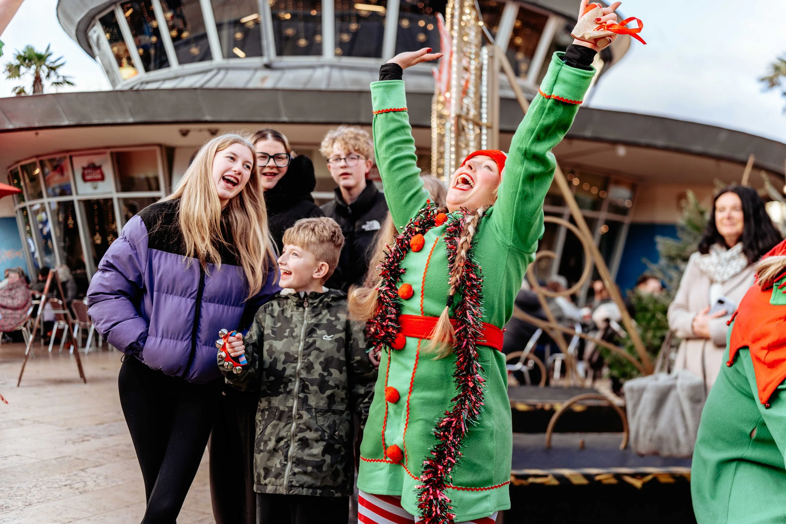 A woman dressed as an elf celebrating on a holiday-themed amusement park ride with children and other adults around her.