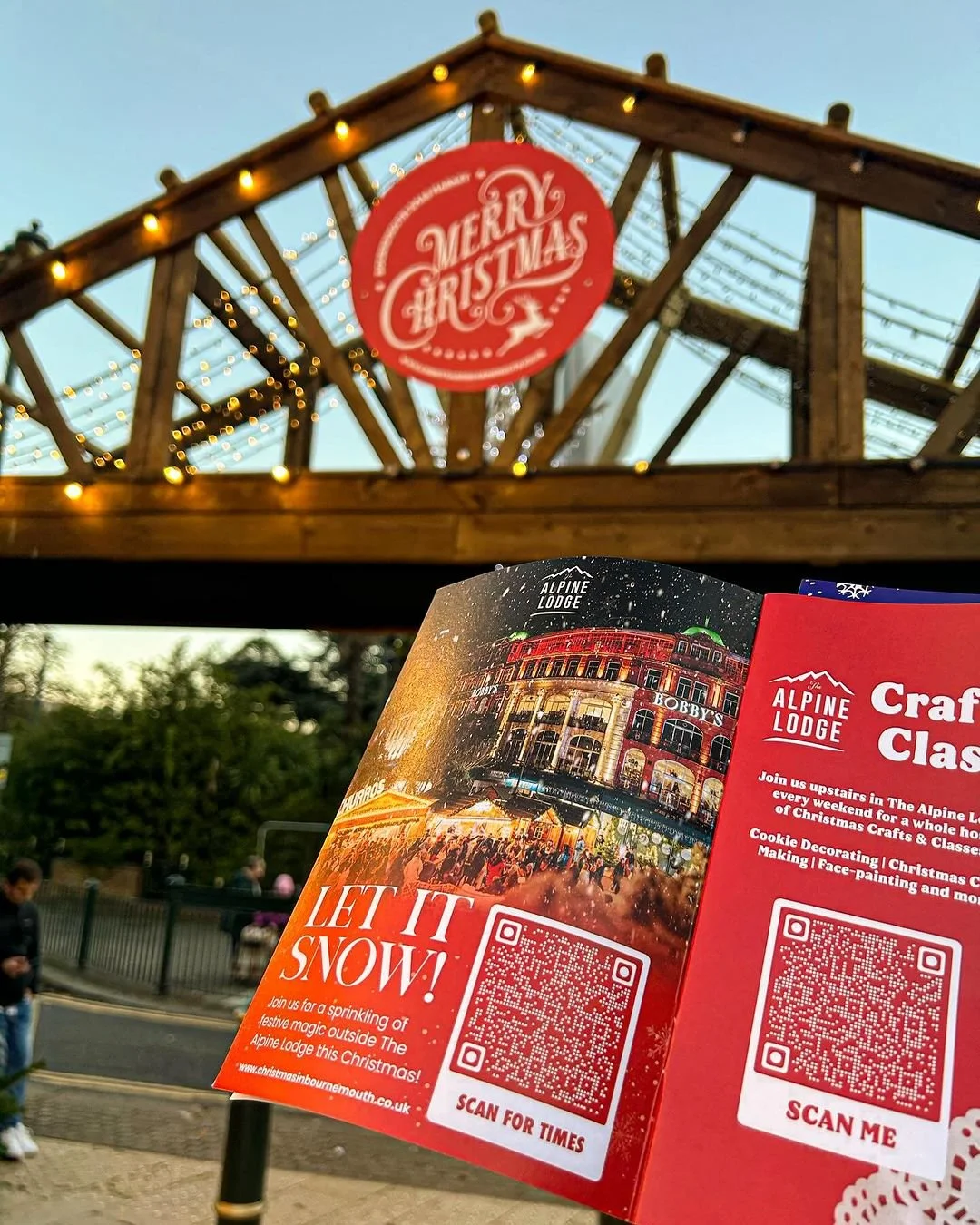 Outdoor scene of a wooden archway decorated with Christmas lights and a red circular sign that says "Merry Christmas". A person is holding a brochure or pamphlet featuring a festive scene with a building illuminated at night, with text promoting holi