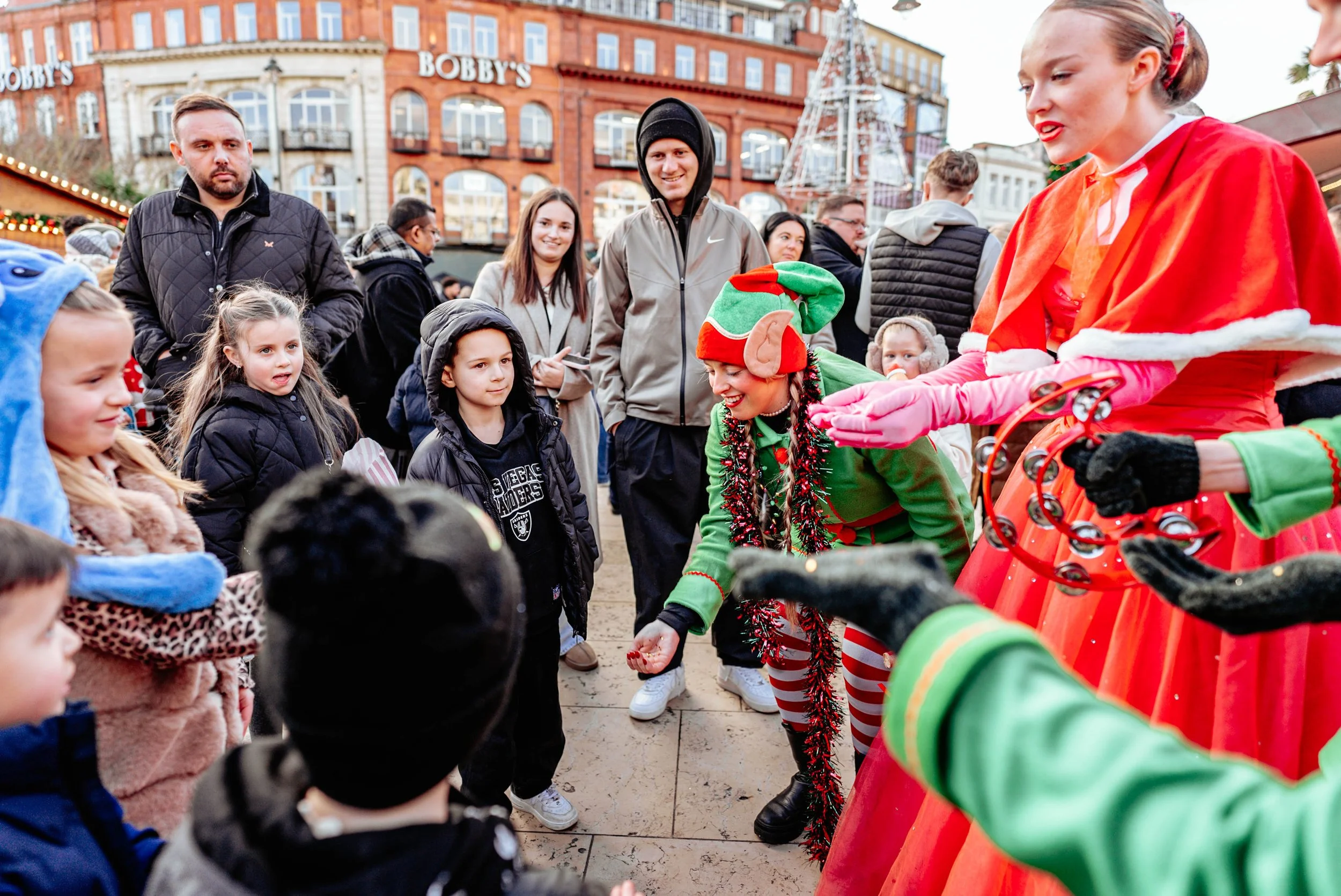 Children and adults gathered at a festive outdoor Christmas event, with entertainers dressed as elves and Santa Claus, during daytime in a city square decorated with holiday lights and holiday buildings in the background.