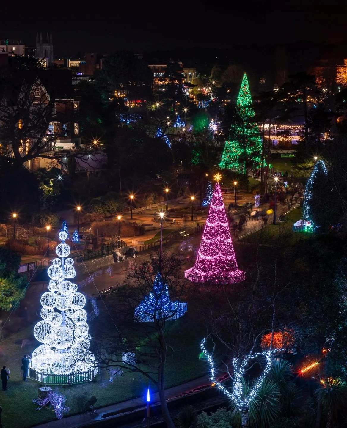 Night scene of a park decorated with colorful illuminated Christmas trees and holiday lights. Several trees are lit with pink, blue, green, and white lights, creating a festive holiday display.