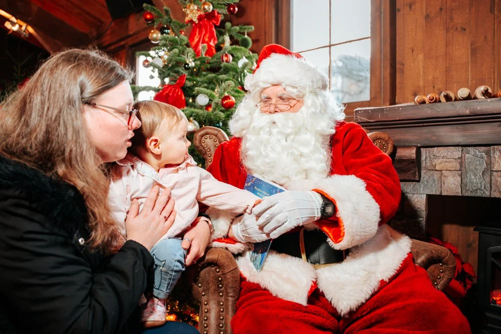 A young child and a woman sitting with Santa Claus in front of a decorated Christmas tree. Santa is holding a gift.