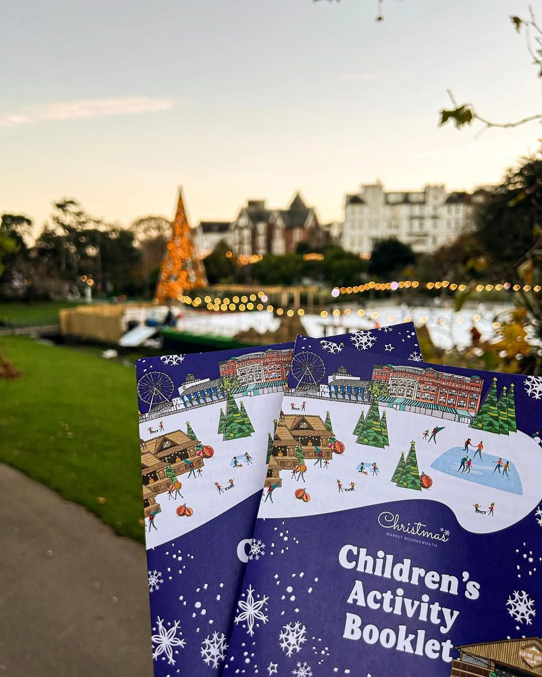 Children's activity booklets with a festive winter scene on the cover, held outdoors near a lakeside park with a Christmas tree and string lights at sunset.