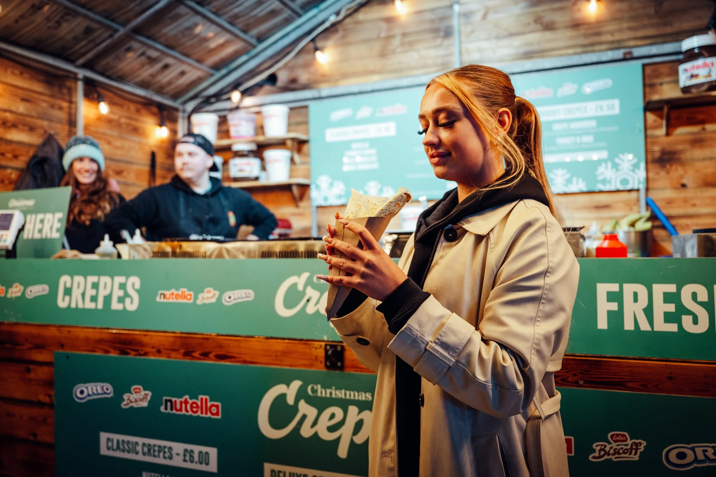 A woman holding a cone of crepes at a food stall with a green sign that reads "Crepes" and branding from Nutella, Oreo, and Bischoff, with two people inside the stall in the background.