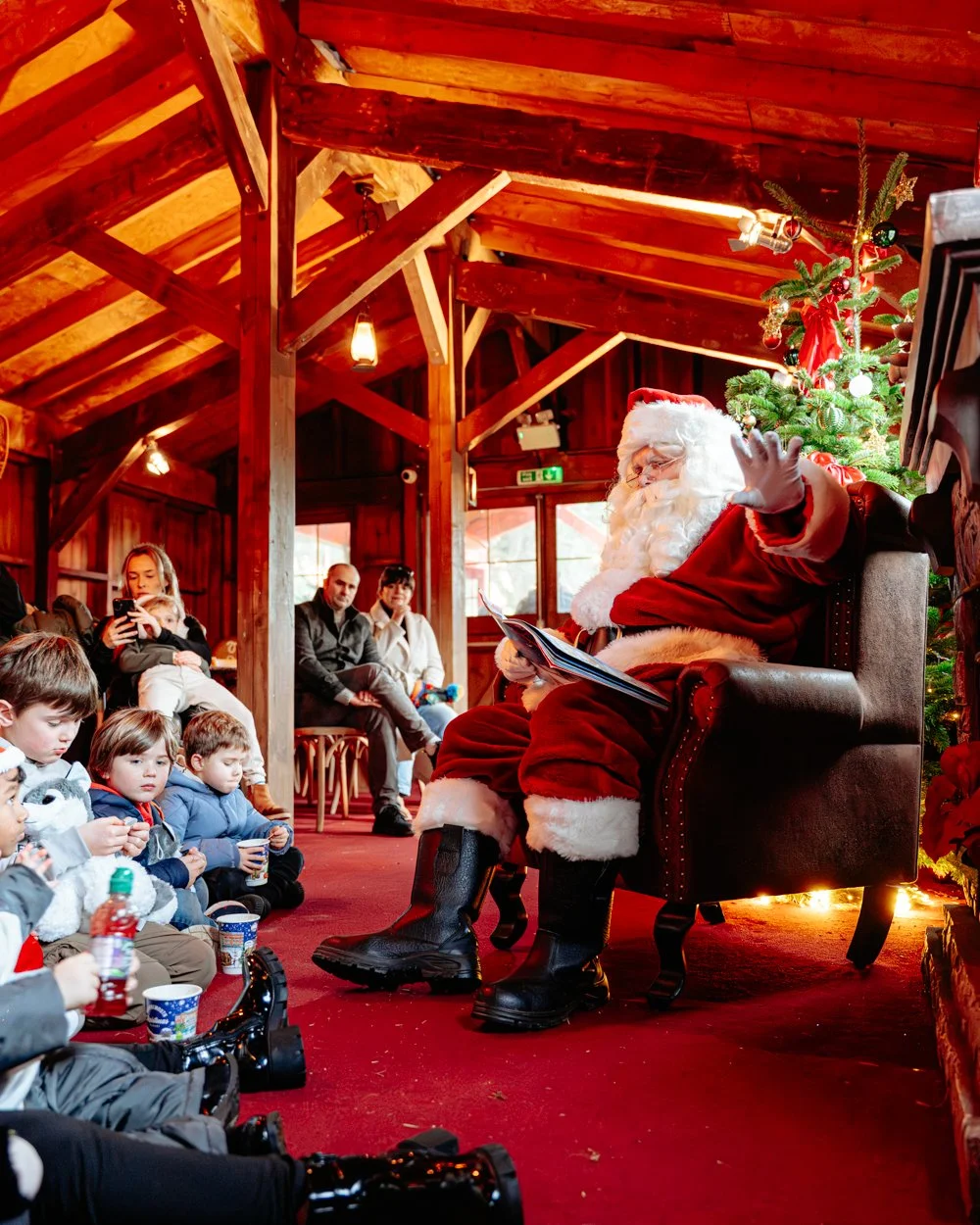 Santa Claus with a white beard, wearing a red suit and black boots, sitting on a brown armchair next to a decorated Christmas tree, raising his hand in a greeting or wave to children seated on the floor during a holiday event.