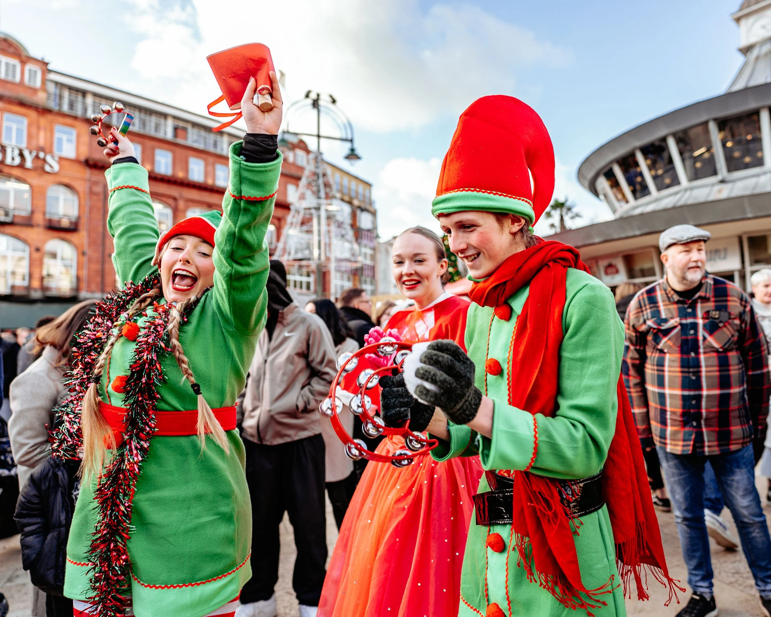 People celebrating outdoors during a festive event, dressed in Christmas-themed costumes, with buildings in the background.