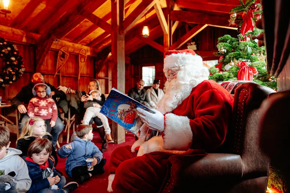 Santa Claus reading a book to children during Christmas in a decorated wooden room with a Christmas tree.