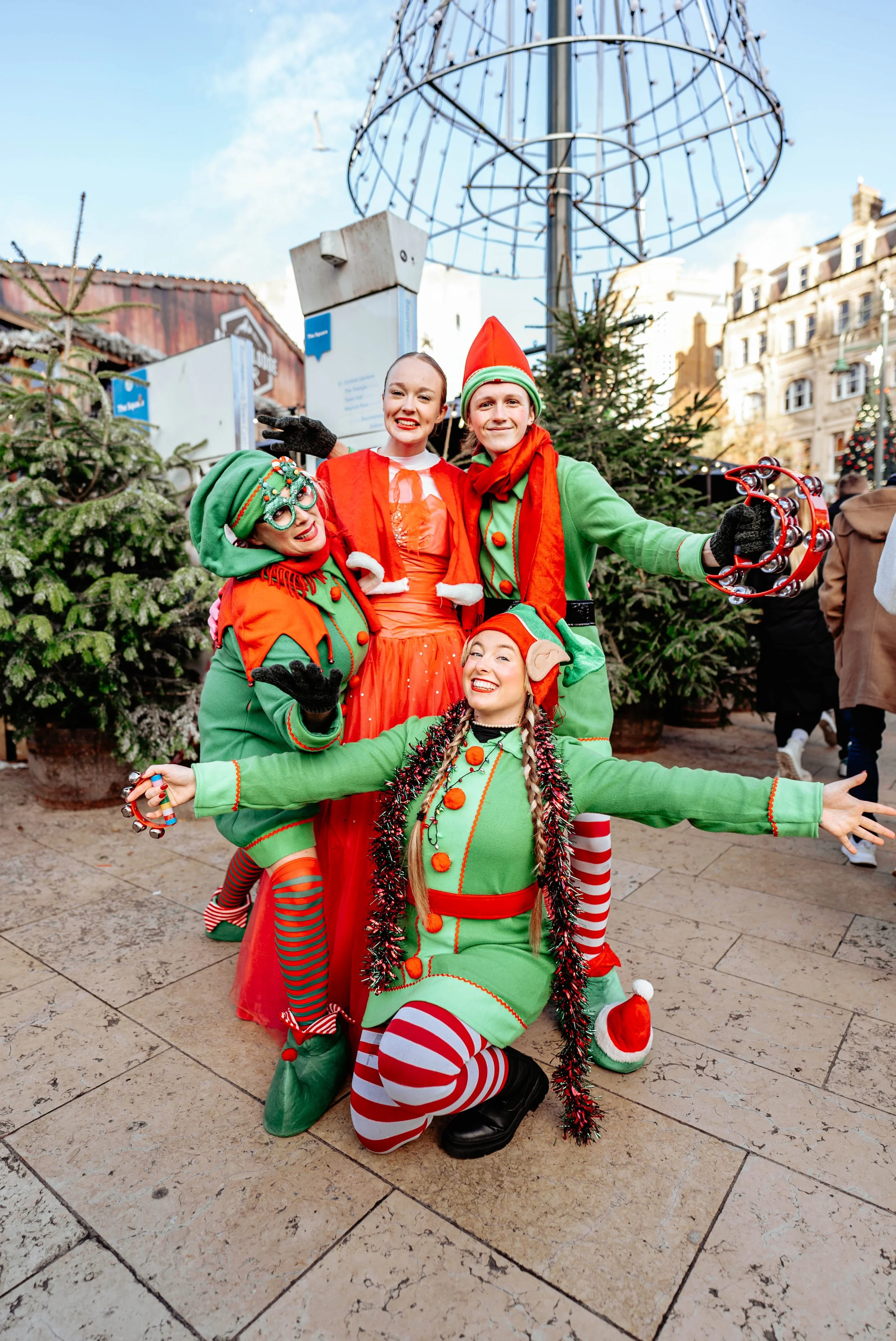 Four people dressed as Christmas elves and a Christmas fairy posing together outdoors during the holiday season in front of Christmas trees and a cityscape.