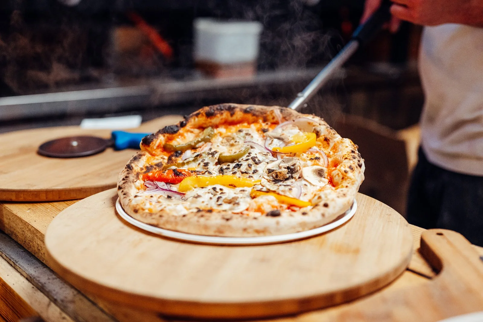 A freshly baked pizza with assorted toppings including yellow and red peppers, mushrooms, and onions on a wooden serving board.