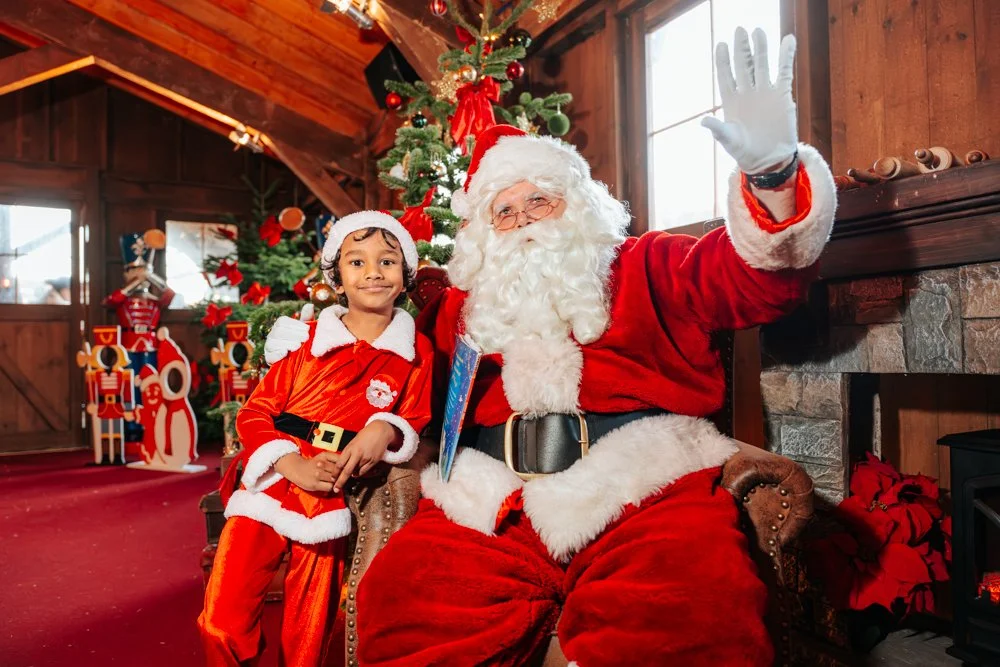 A young girl dressed as Santa Claus sitting next to a man dressed as Santa Claus in a festive, Christmas-decorated room, with a Christmas tree and holiday decorations in the background, inside a cozy wooden cabin.