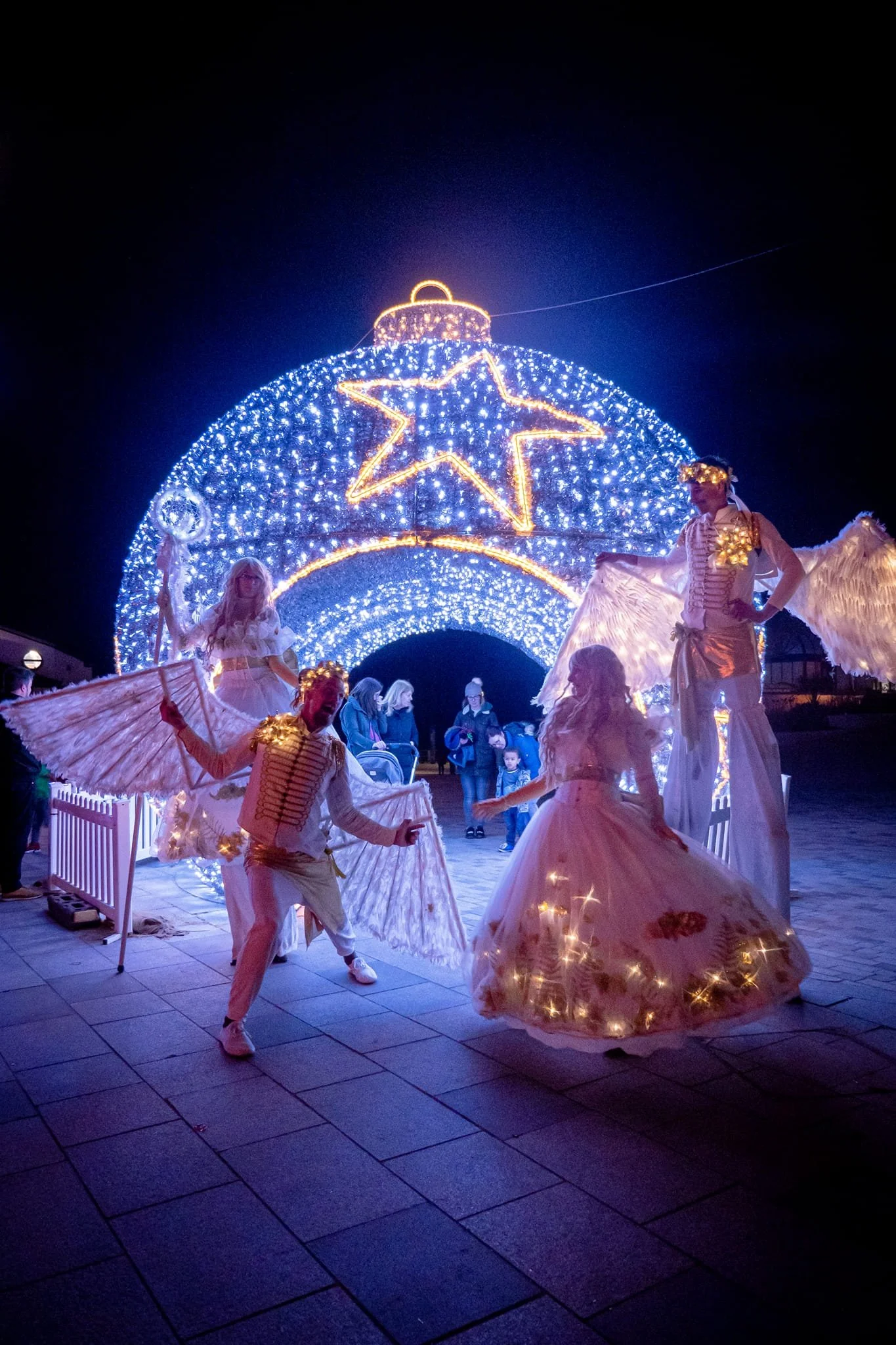 People dressed as angels performing in front of a large, illuminated Christmas display with a star and ornaments at night.