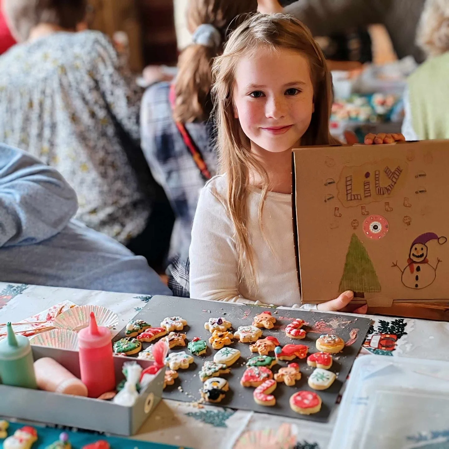 A young girl with long red hair and a white shirt sitting at a table with decorated Christmas cookies and frosting supplies. She is holding a brown gift box decorated with a snowman and holiday drawings, smiling at the camera.