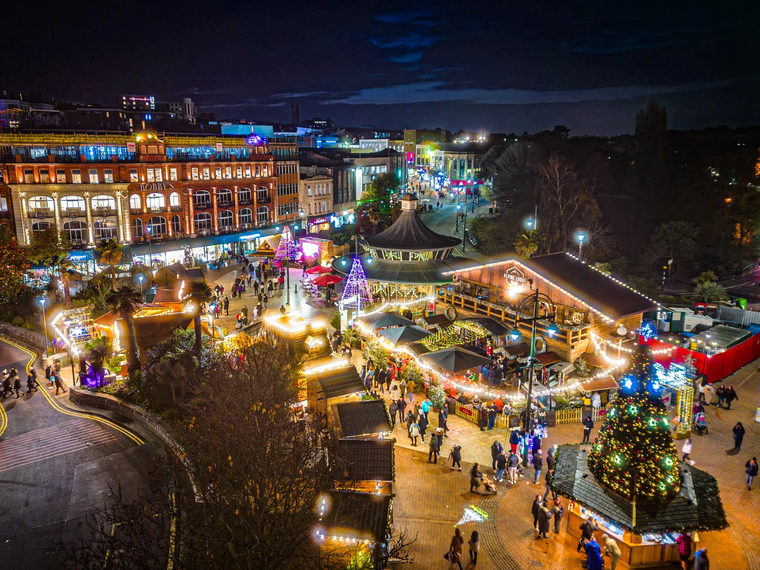 Nighttime view of a festive outdoor area decorated with Christmas lights, trees, and market stalls. Crowds of people are walking and shopping, with a large, illuminated Christmas tree in the foreground.