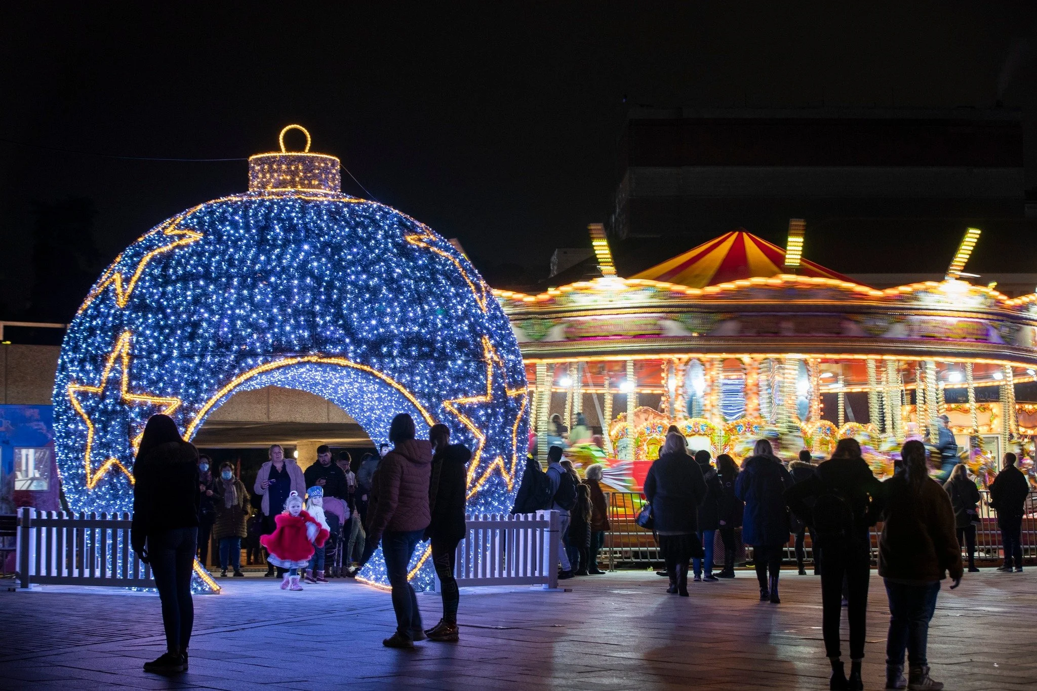 Night scene at a carnival with a large illuminated blue Christmas ornament with yellow star patterns and a carousel with bright, colorful lights and a striped red and yellow top.