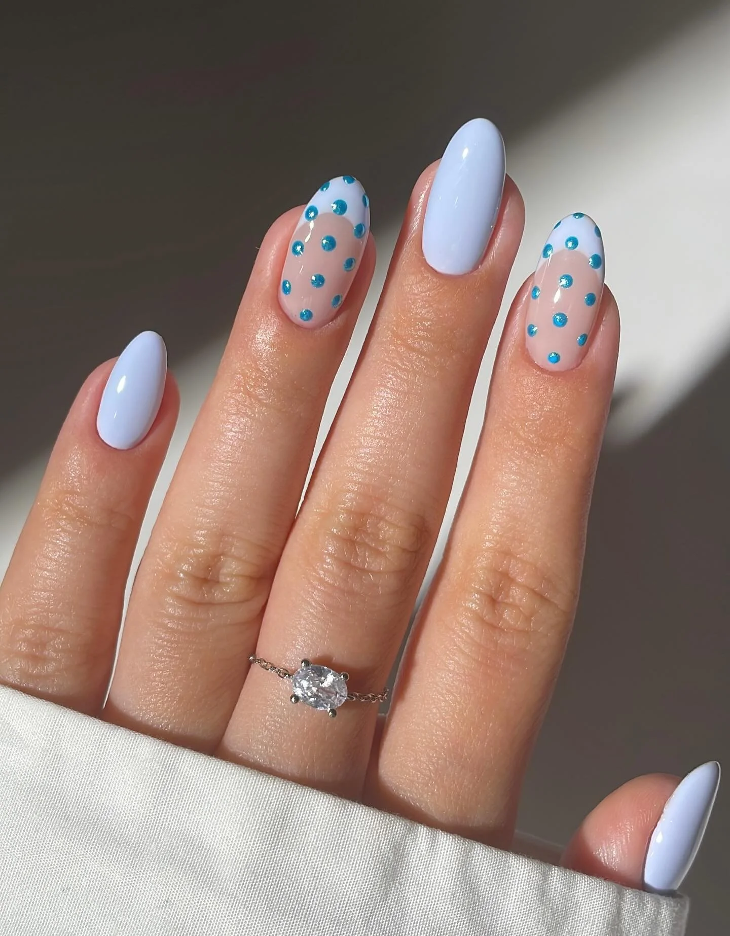 Hand with manicured nails featuring light blue and clear polish with blue dot accents, wearing a silver ring with a large clear gemstone.