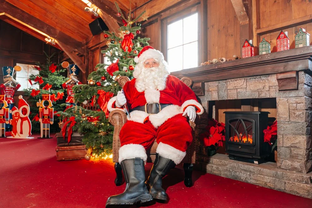 Santa Claus sitting in a cozy wooden room with a fireplace and decorated Christmas tree, surrounded by holiday decorations.