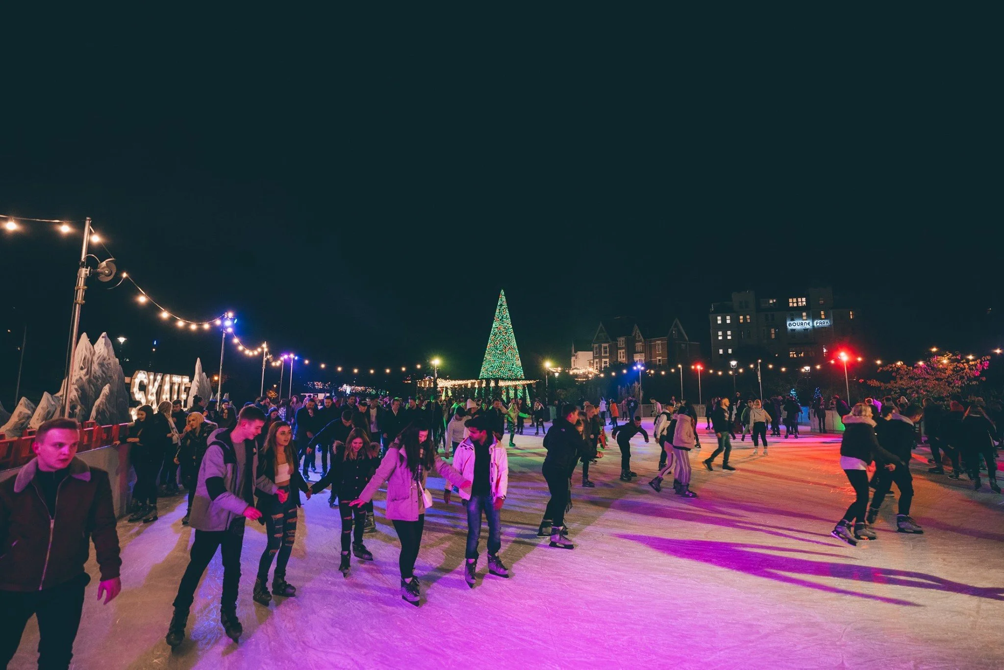 People ice skating at an outdoor rink decorated with a large Christmas tree and festive lights at night.