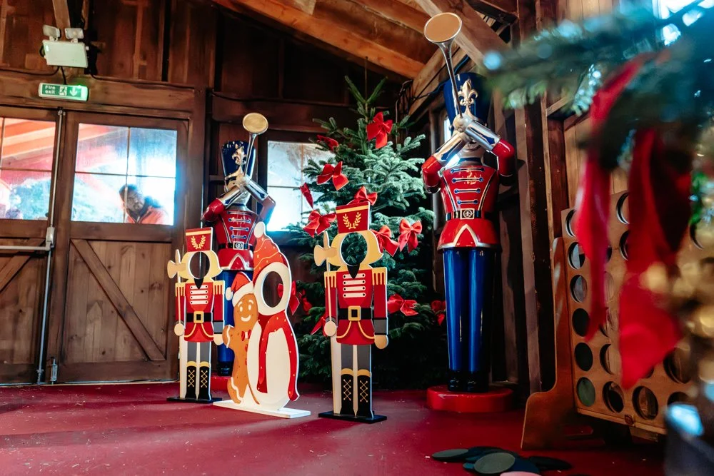 Decorative Christmas scene with a small Christmas tree adorned with red bows and ornaments, surrounded by three large wooden nutcracker figures in red uniforms, one holding a drum, inside a wooden building with a red carpeted floor.