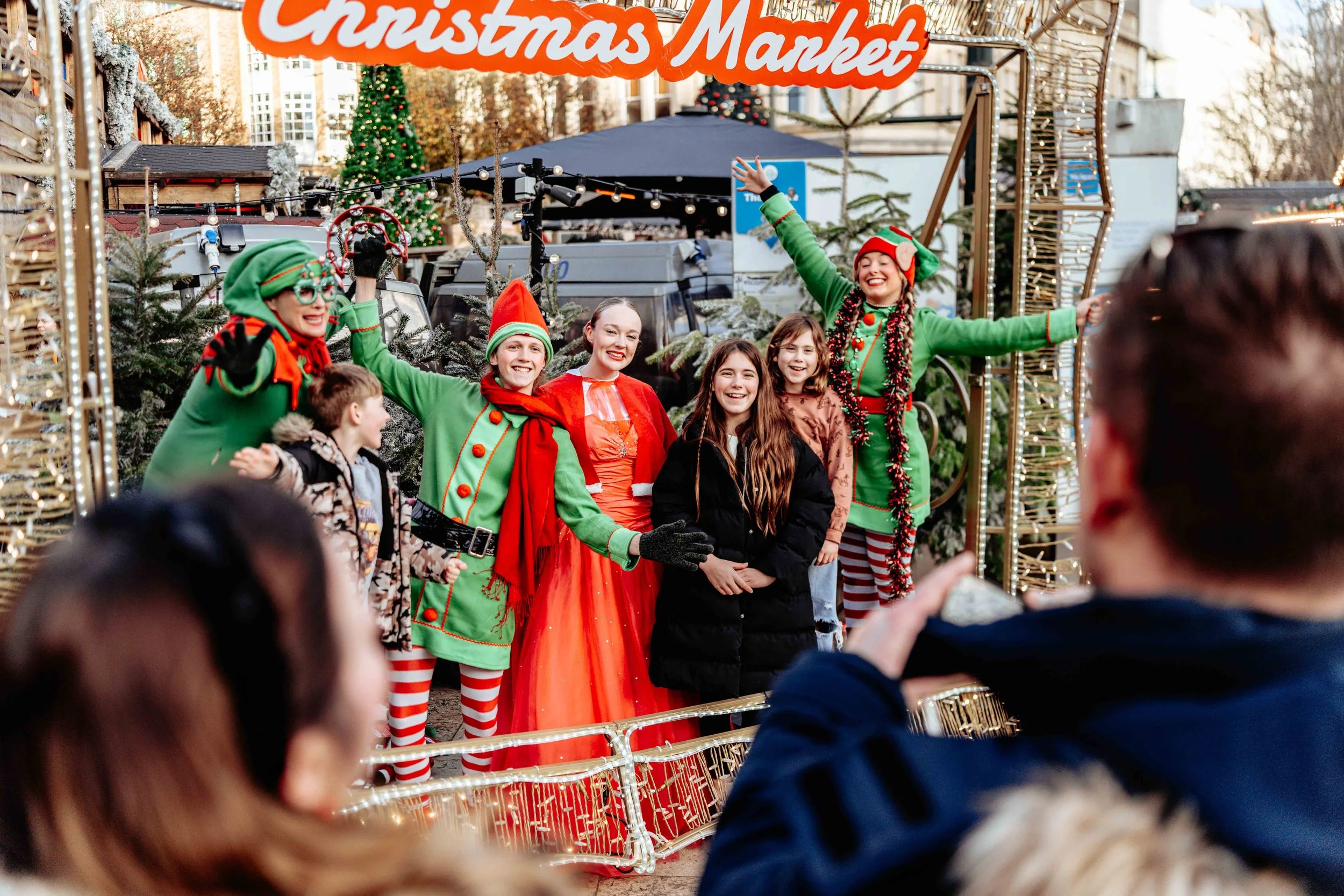 People dressed as Christmas elves and a woman in a holiday costume posing for photos at a Christmas market with Christmas trees and festive lights.