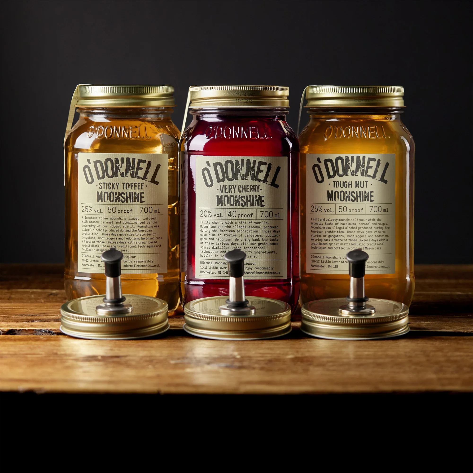 Three jars of Ó'Donnell Moonshine in different flavors—Sticky Toffee, Very Cherry, and Tough Nut—on a wooden surface with their lids separately placed in front of each jar.