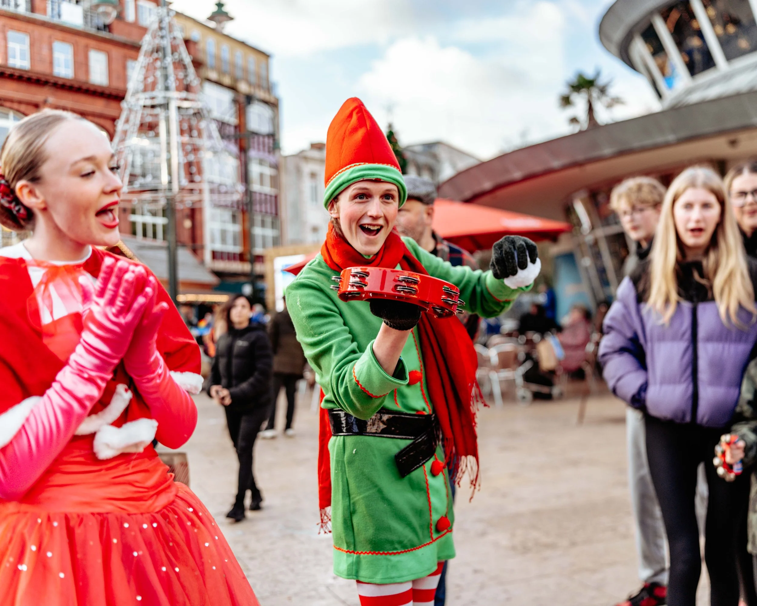People dressed in festive holiday costumes, including an elf holding a red tambourine, standing outdoors at a holiday market or street fair with decorations and other people in the background.