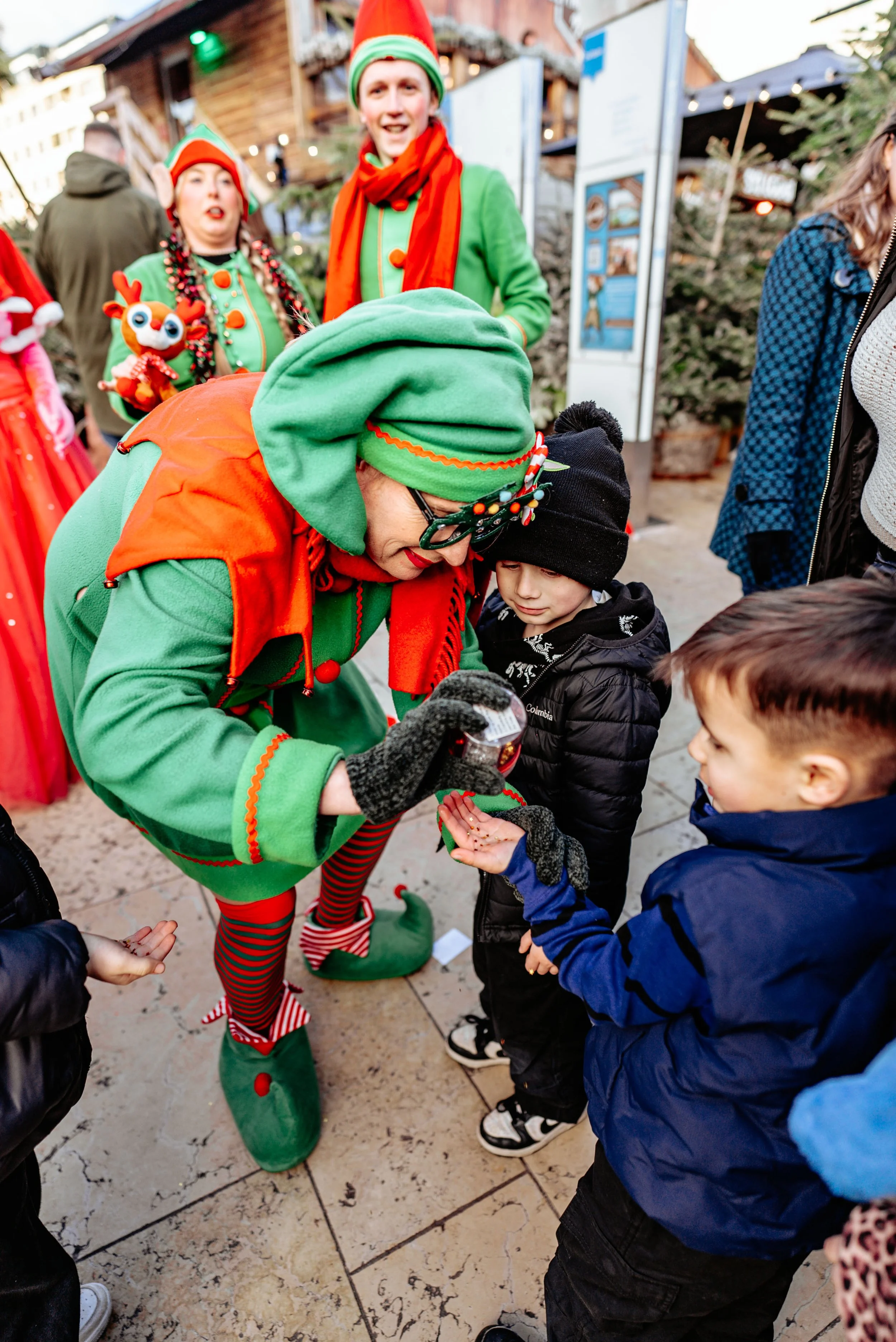 A woman dressed as an elf giving out small items to children at a festive outdoor event, with other people celebrating in the background.