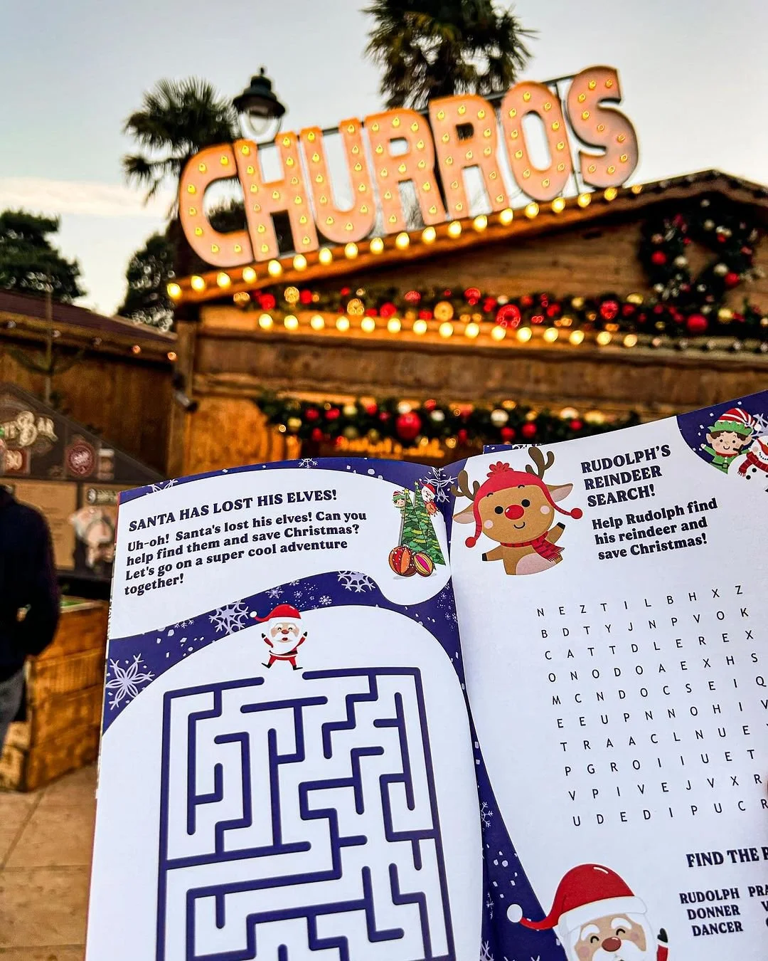 A festive Christmas scene with a large illuminated 'CHAROS' sign in the background, decorated with Christmas wreaths and lights, and a holiday activity booklet with a maze and word search in the foreground.