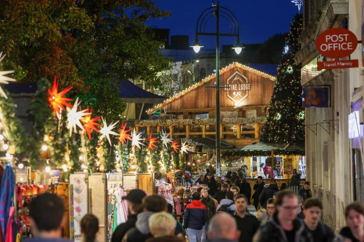 A crowded Christmas market street decorated with red and white star-shaped lights, green garlands, and Christmas trees. A wooden building with a sign that reads 'Alpine Lodge Bar' is in the background, illuminated with string lights. People are shopping and socializing in the evening.