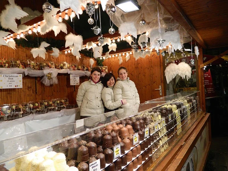 Three women in cream-colored coats standing behind a chocolate stall decorated with white angel wings and Christmas ornaments at a festive market.