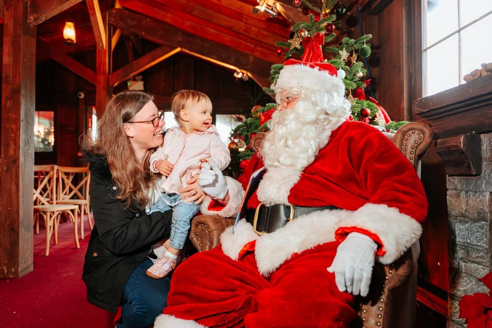 A woman with glasses and long hair sitting with a young girl on her lap, facing Santa Claus in a cozy, decorated Christmas setting with a Christmas tree. Santa is dressed in his traditional red suit with white fur trim, wearing glasses, and sitting i