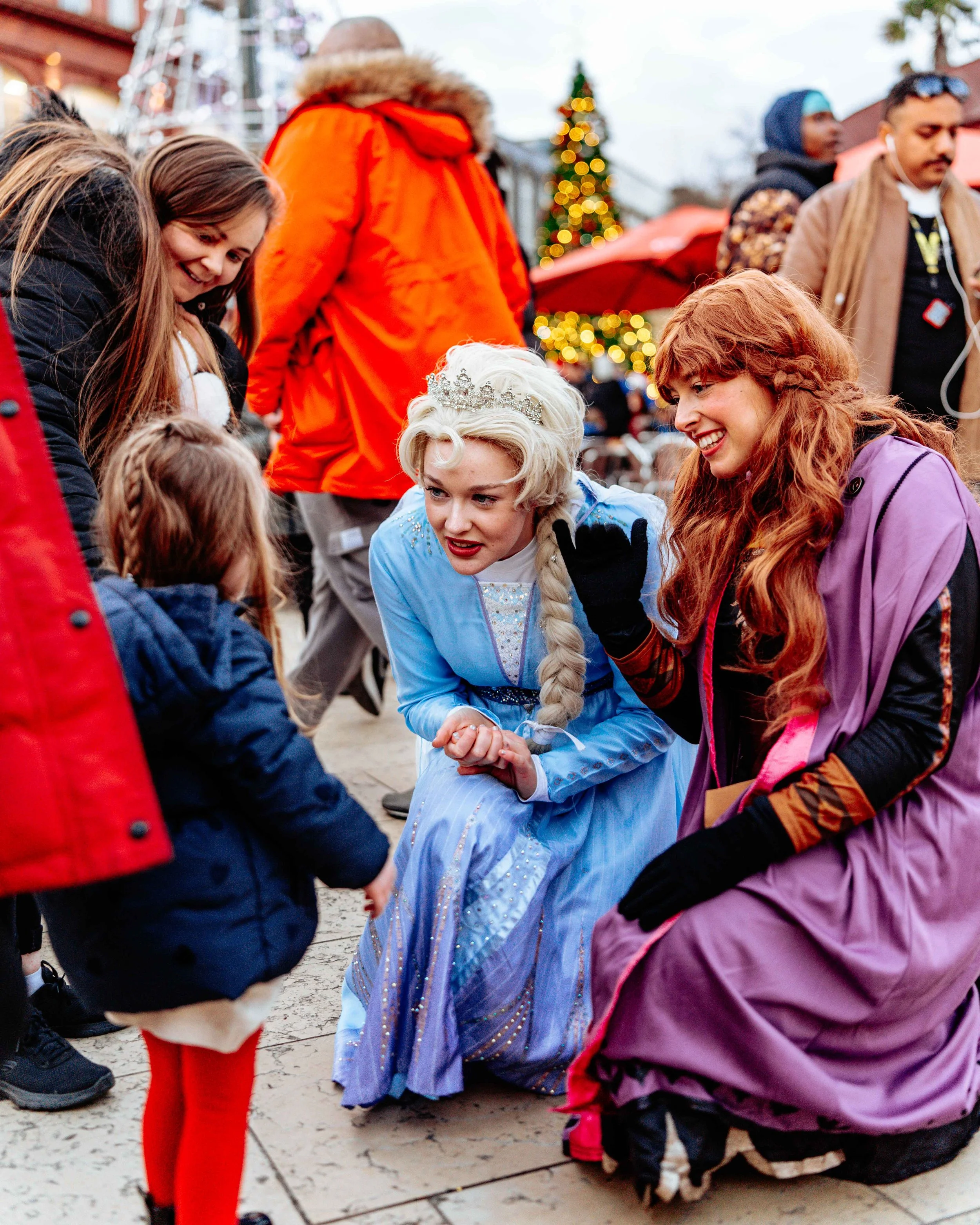 A woman dressed as Elsa from Frozen, wearing a blue gown and a tiara, is talking to a young girl and her family at a festive outdoor market decorated with Christmas lights and a Christmas tree.