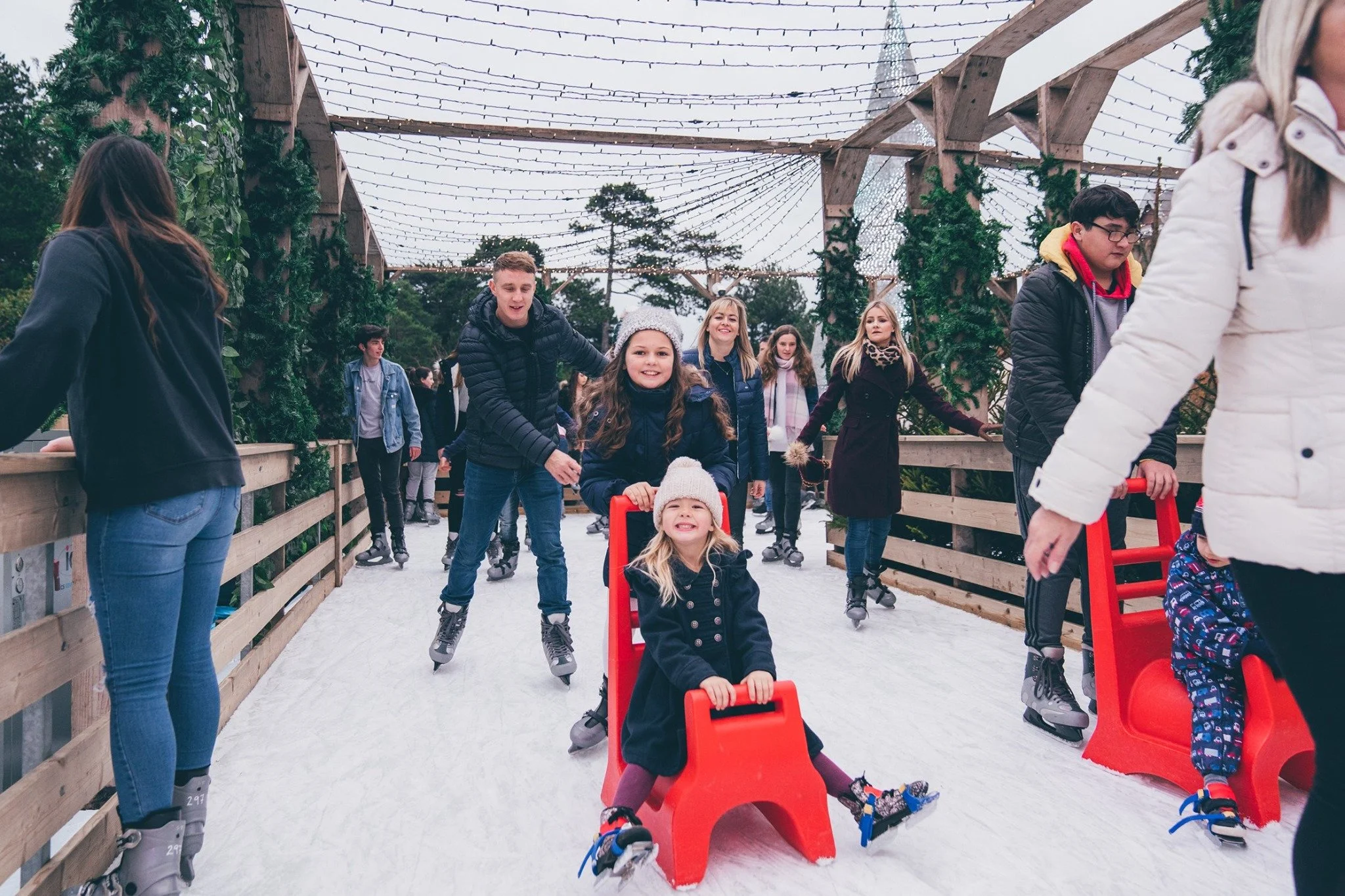 People enjoying ice skating at an outdoor rink decorated with string lights and greenery, with a young girl sitting on a red sled on the ice.