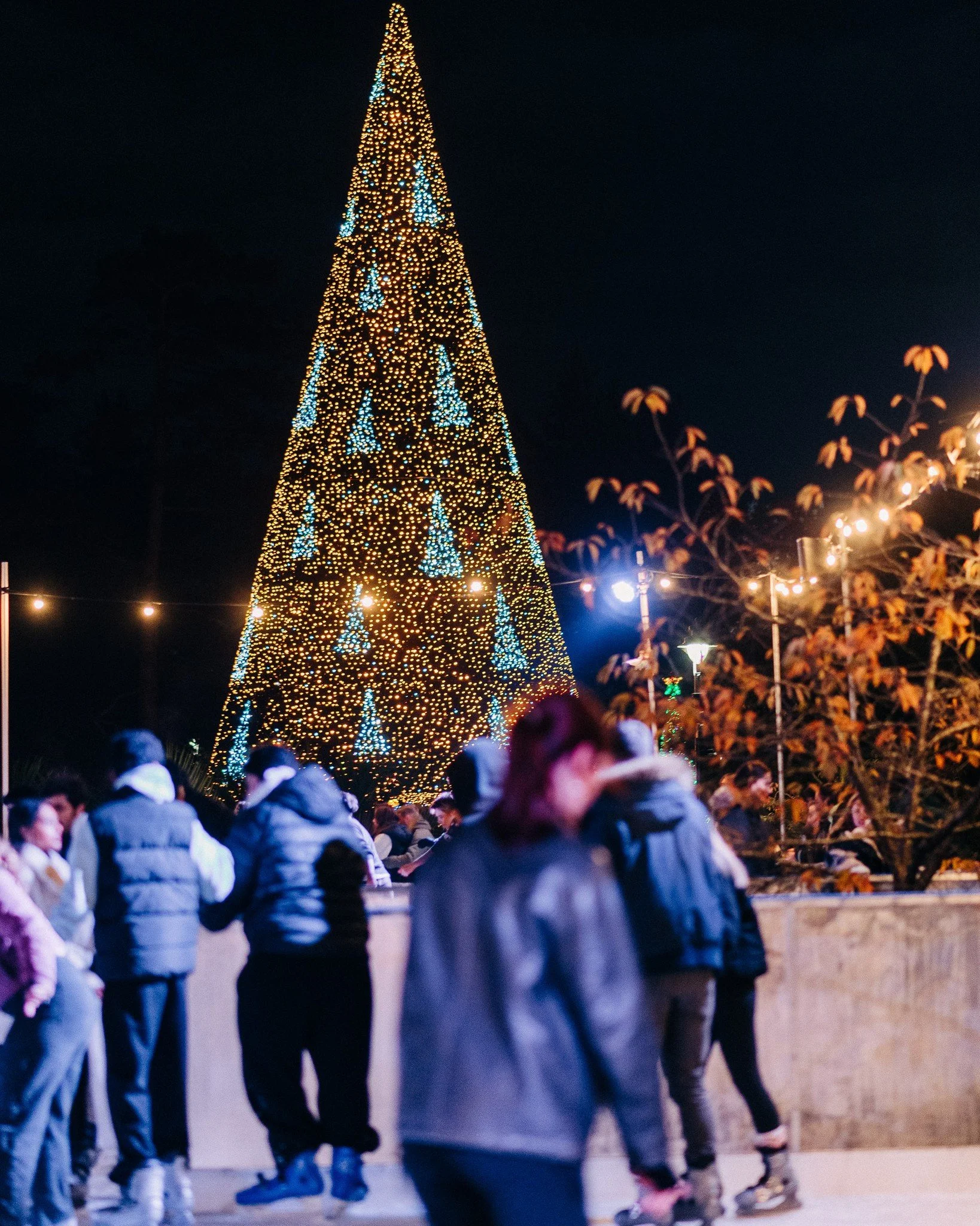 People ice skating outside at night with a large decorated Christmas tree in the background.