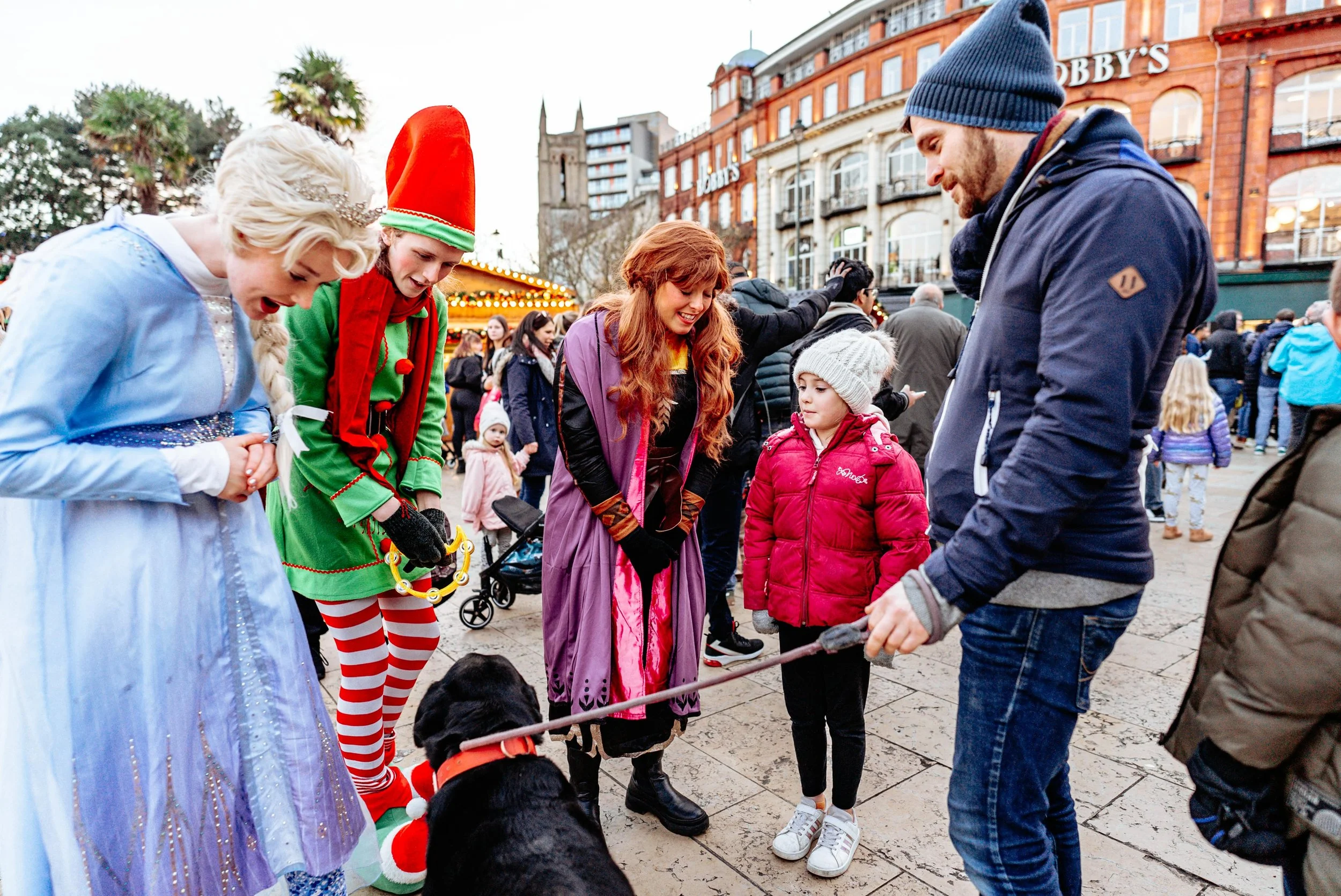 People dressed in Christmas costumes and winter clothing, gathered around a black dog on a city square during a festive event.
