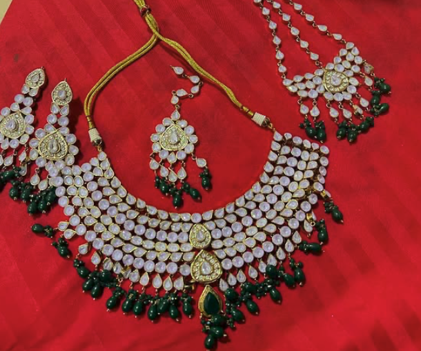 Elegant traditional jewelry set including a necklace and matching earrings, adorned with white gemstones and green beads, displayed on a red fabric background.