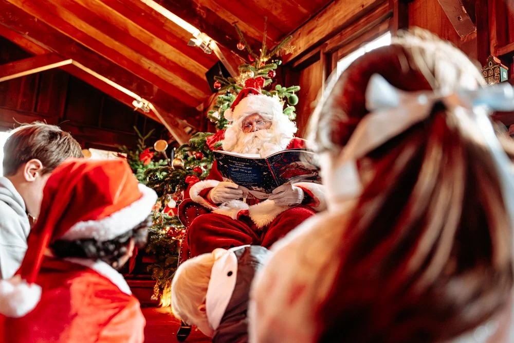 Santa Claus reading a book to children in Christmas hats in front of a decorated Christmas tree in a cozy wooden room.