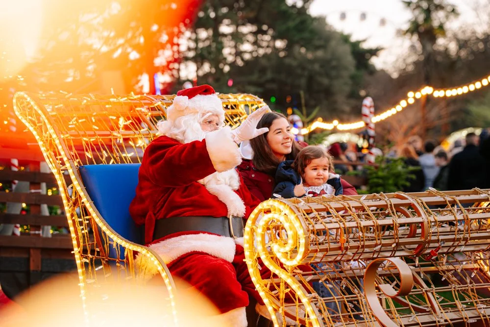 Santa Claus, a woman, and a young girl sitting on a festive sleigh with lights at an outdoor Christmas event.