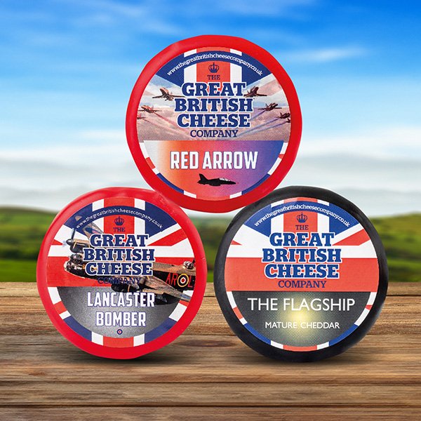 Three rounds of British cheese from The Great British Cheese Company on a wooden surface with a grassy field and blue sky in the background.